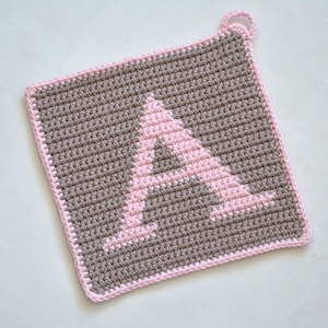 Op de afbeelding: Een bruine en roze gehaakte ovenwant met een roze letter A in het midden.