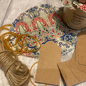 May include: A collection of craft supplies including a jar of unscented bee wax candle, patterned fabric circles, brown tags, rubber bands, and twine. The fabric has a red, blue, and green floral design.