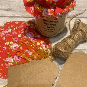 May include: A small glass jar candle with a red floral fabric lid. The jar contains unscented beeswax and is labeled "Hand Poured 130 grams". Also shown are brown tags, twine, and a piece of red floral fabric.