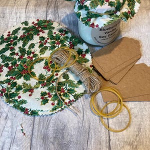 May include: A white soy candle with a holly-patterned fabric lid. The candle is labelled "Blueberry Vanilla Soy Candle" and "Hand Poured". The image also shows a holly-patterned fabric circle, twine, rubber bands, and brown paper gift tags.