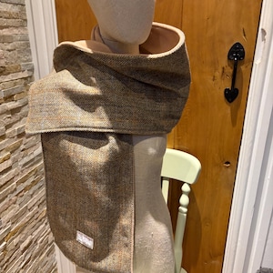 May include: A brown and tan tweed scarf with a herringbone pattern is draped on a mannequin. The scarf has a wide collar and a long, rectangular front panel. The background includes a wooden door and a light green chair.