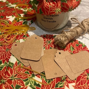 May include: A glass jar candle with a red and green fabric cover. The candle label reads "Nut & Pecan Danish Soy Candle" and "Country Crafts". The candle is surrounded by brown gift tags, rubber bands, and twine.