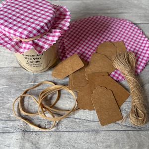 Peut inclure: Un pot en verre de bougie à la cire d'abeille non parfumée avec une housse en tissu vichy rose et blanc, accompagné de plusieurs étiquettes cadeaux en papier brun, de ficelle de jute, d'élastiques et d'un tissu vichy rose circulaire.