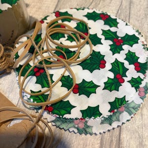 May include: A collection of circular fabric food covers, patterned with green holly leaves and red berries on a white background. Several elastic bands and twine are next to the covers. A jar with a holly-patterned lid is visible.