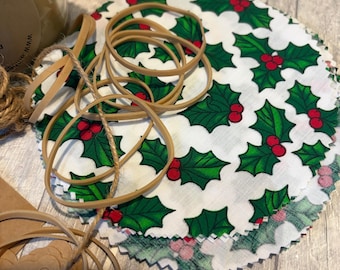 May include: A stack of round, white fabric food covers with a green holly and red berry pattern. Several tan rubber bands and a length of twine are next to the covers. A jar with a holly-patterned lid is visible in the background.