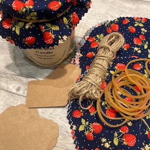 May include: A jar candle with a navy blue strawberry-patterned lid and fabric wrap. The image also shows fabric circles, twine, rubber bands, and blank tags, indicating a craft or DIY project.