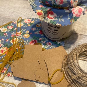 May include: A blue floral fabric-covered jar containing a bee wax candle. The jar has a label that reads "Bee Wax Candle Hand Poured 130 grams" and a website address. The image also shows brown paper gift tags, rubber bands, and twine.