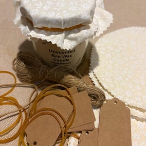 May include: A small amber candle jar with a floral fabric lid, labelled "Unscented Bee Wax Candle." The image includes twine, brown tags, and rubber bands. The candle is hand-poured, suggesting a natural product.