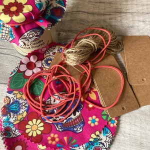 May include: A stack of pink fabric circles with a sugar skull and floral pattern. The fabric circles are tied together with a red rubber band. There is a jar with a pink and white fabric lid, a ball of twine, and two brown paper tags in the background.