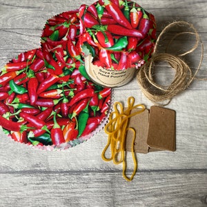 May include: A set of three red and green chilli pepper patterned fabric jar covers with a brown twine roll, rubber bands, and two brown paper tags. The jar cover is labelled "Peppercorn Soya Candle Hand Poured".