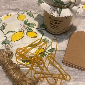 以下が含まれることがあります： レモンと花の模様の布の蓋が付いた、無香料の蜜蝋キャンドルの瓶。画像には、お揃いの布の正方形、輪ゴム、紐、茶色の紙タグも含まれています。キャンドルは手作業で注がれ、重さは130グラムです。