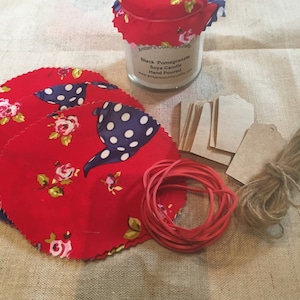 May include: A red and white fabric covered jar with a black and white polka dot teapot design. The jar contains a soy candle with the text "Black Pomegranate Soy Candle Hand Poured" and the website address "www.amberscountrycrafts.com". The image also shows red fabric circles, brown paper tags, a red rubber band, and brown twine.