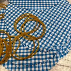 May include: Close-up of blue and white gingham fabric circles with a scalloped edge. Several amber-coloured rubber bands are scattered across the fabric. The fabric is arranged on a light-coloured wooden surface.