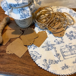 May include: A candle jar with a blue and white toile fabric lid cover, accompanied by several brown paper tags, a bundle of rubber bands, a length of twine, and a circular piece of matching blue and white toile fabric with pinked edges.