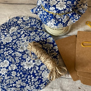 May include: A blue and white floral fabric jar lid with an elastic band around the top. The jar lid is sitting on a wooden surface with a piece of twine and a stack of brown paper cards.