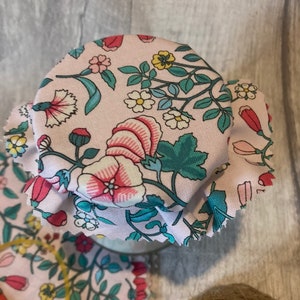 Peut inclure: Un couvercle de pot en tissu rose avec un motif floral dans des tons de rose, vert, bleu et jaune. Le tissu est froncé autour du bord du couvercle.
