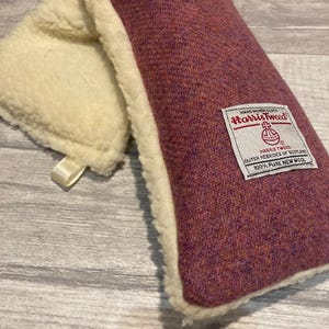 May include: A rectangular, burgundy-colored Harris Tweed pillow with a cream-colored, fluffy border. The pillow has a label that reads "Harris Tweed" and "100% Pure New Wool". The pillow is on a light gray wooden surface.