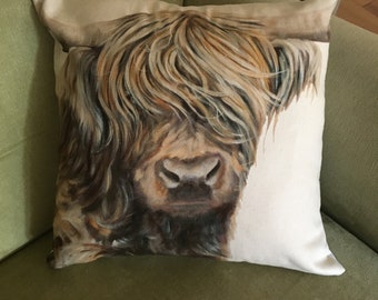 May include: A brown and white Highland cow with long, shaggy hair is featured on a cream-colored throw pillow. The pillow is sitting on a green couch.