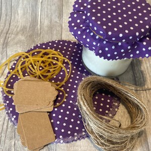 May include: A glass jar with a purple polka dot fabric lid and coaster. The image also includes a bundle of brown twine, a stack of brown tags, and yellow rubber bands. The items are arranged on a grey wooden surface.