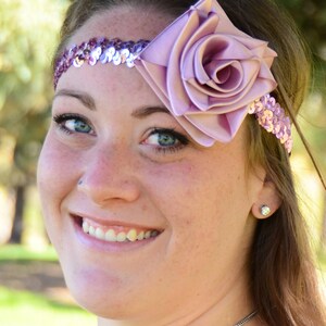 Pode incluir: Uma mulher usando uma faixa de cabeça com uma grande rosa de cetim lilás claro e uma fileira de lantejoulas iridescentes. A faixa é usada na testa.