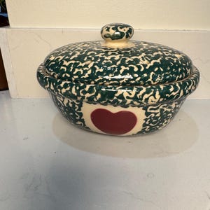 May include: A ceramic serving bowl with a lid, featuring a cream base and a dark green speckled pattern. The bowl has a red heart design on the front. The lid has a small knob. This decorative bowl is ideal for serving food, and is a kitchen essential.