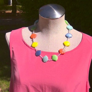 May include: A pink sleeveless dress with a colorful seashell necklace. The necklace has yellow, blue, green, and red seashells.