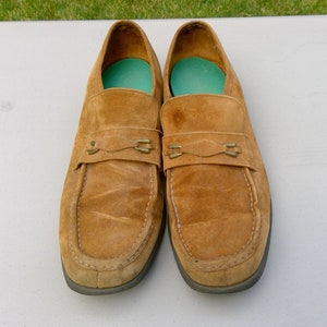May include: A pair of brown suede loafers with gold buckles. The loafers have a slightly rounded toe and a low heel.