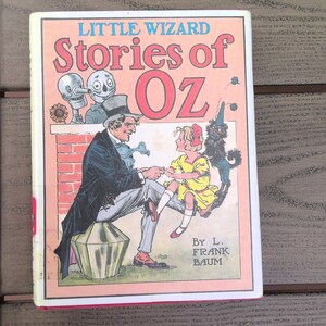 May include: A vintage book cover with a pink background and the title "Little Wizard Stories of Oz" in red and blue lettering. The cover features illustrations of a man in a top hat, a girl in a yellow dress, and a black dog.