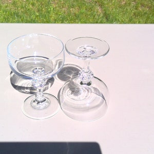May include: Two clear champagne glasses with ornate stems are displayed on a light-colored surface. One glass is upright, and the other is inverted, casting shadows. The glasses are made of transparent glass.
