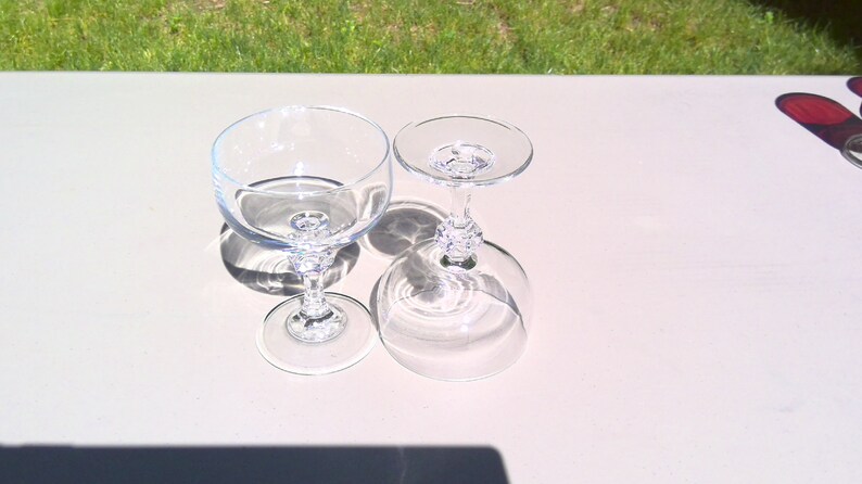 May include: Two clear champagne glasses with ornate stems are displayed on a light-colored surface. One glass is upright, and the other is inverted, casting shadows. The glasses are made of transparent glass.