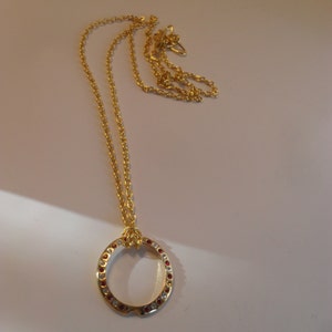 May include: A gold chain necklace with a circular pendant. The pendant is adorned with small red and clear rhinestones.