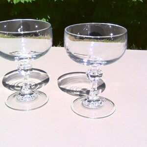 May include: Two clear, stemmed champagne glasses with a round bowl and a decorative stem. The glasses are made of clear glass and are sitting on a white surface. The glasses cast a shadow on the surface.