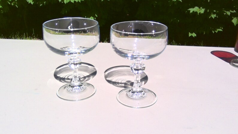 May include: Two clear, stemmed champagne glasses with a round bowl and a decorative stem. The glasses are made of clear glass and are sitting on a white surface. The glasses cast a shadow on the surface.