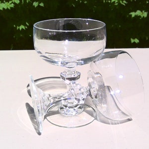 May include: Three clear, stemmed champagne glasses. One glass is upright, while two are lying on their sides. The glasses have a round bowl and a decorative, faceted stem. The background is a blurred view of green foliage.
