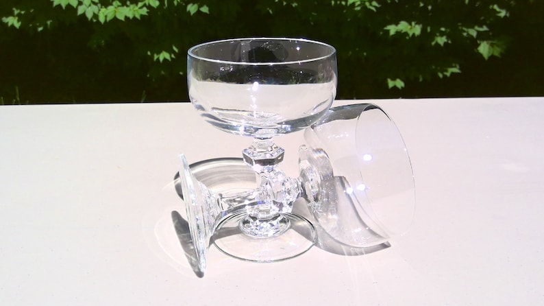 May include: Three clear, stemmed champagne glasses. One glass is upright, while two are lying on their sides. The glasses have a round bowl and a decorative, faceted stem. The background is a blurred view of green foliage.