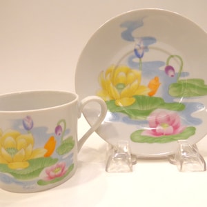 May include: A white teacup and saucer set with a floral design. The design features yellow, pink, and purple water lilies with green leaves on a blue background.