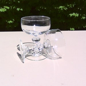 May include: Three clear crystal champagne coupe glasses. One glass is upright, while the other two are lying on their sides. The glasses have a round bowl and a decorative, faceted stem. The background is a blurred view of green foliage.