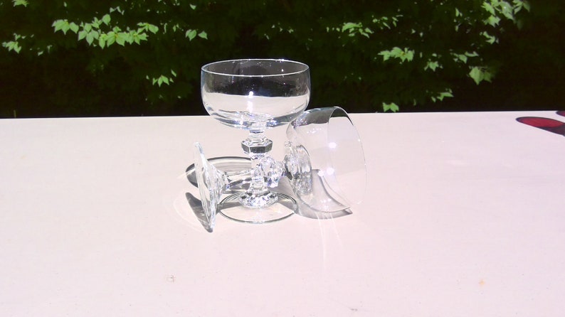 May include: Three clear crystal champagne coupe glasses. One glass is upright, while the other two are lying on their sides. The glasses have a round bowl and a decorative, faceted stem. The background is a blurred view of green foliage.