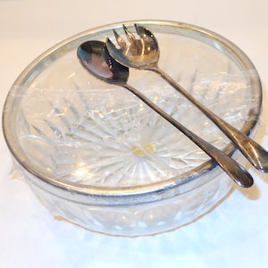 May include: A clear glass serving bowl with a silver rim, containing a spoon and a fork. The bowl is wrapped in plastic. The spoon and fork are silver and appear to be serving utensils.