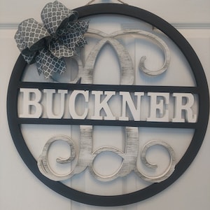May include: A black and white wooden door hanger with the name "BUCKNER" in white letters and a large white letter "B" with a decorative flourish. The hanger is decorated with a gray and white bow.