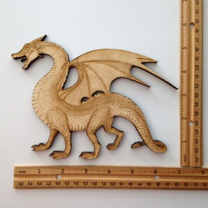 May include: A wooden dragon cutout, light brown in colour. The dragon is standing on four legs with its wings spread out. The dragon is approximately 15 cm long.