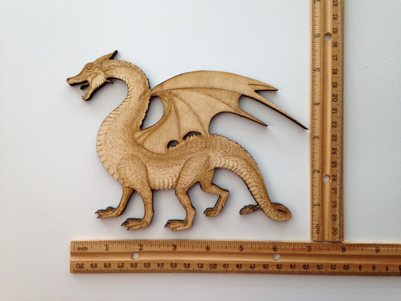 May include: A wooden dragon cutout, light brown in colour. The dragon is standing on four legs with its wings spread out. The dragon is approximately 15 cm long.
