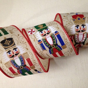 May include: A beige ribbon with a red trim features a repeating pattern of nutcracker soldiers in various poses. The nutcrackers are wearing colorful uniforms with gold accents and have detailed facial features.