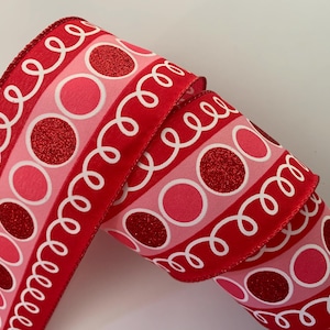 May include: Red and pink decorative ribbon with a repeating pattern of glittery red circles and pink circles, separated by white swirly lines. The ribbon is rolled up, showing the pattern on the front and sides.