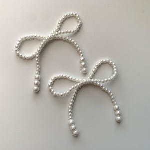 May include: Two white pearl bow-shaped hair accessories. Each bow is made of small, round pearls, with a larger pearl at the end of each strand. The bows are arranged on a white surface.
