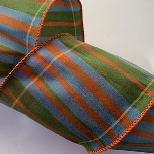 May include: A wide fabric ribbon featuring a plaid pattern with vertical and horizontal stripes in shades of olive green, slate blue, and burnt orange. The edges of the ribbon are finished with a decorative burnt orange stitched border.