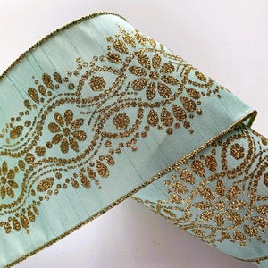 May include: A wide, light mint green ribbon featuring a textured, shimmering gold glitter pattern of repeating floral and paisley-inspired motifs. The edges of the ribbon are finished with a thin, matching gold glitter trim along both sides.