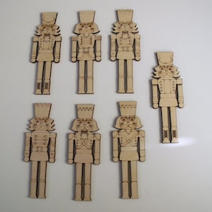 May include: Seven wooden nutcracker ornaments in various poses. Each ornament is painted in a light brown color and features a detailed design of a nutcracker with a hat, mustache, and facial features. The ornaments are all approximately 6 inches tall.
