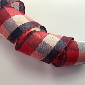 May include: A rolled-up decorative ribbon with a red, white, and navy blue plaid pattern. The ribbon has a stitched red edge and is suitable for crafts, gift wrapping, or holiday decorations.