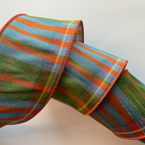 May include: A wide fabric ribbon featuring a plaid pattern in shades of olive green, light blue, and bright orange. The ribbon has a textured, woven appearance and is finished with a distinct, stitched orange edge along both sides of the material.